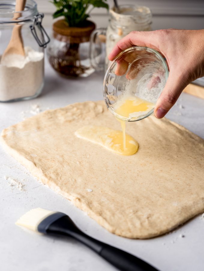 Someone is pouring melted butter onto a rectangle of sourdough cinnamon roll dough.