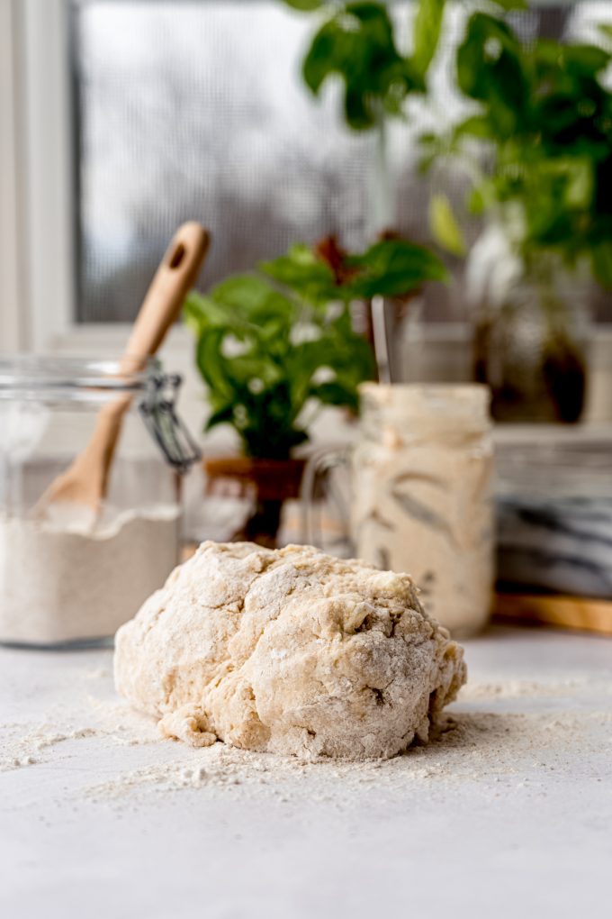 A ball of sourdough starter cinnamon roll dough on a table with a canister of flour and jar of starter in the background.