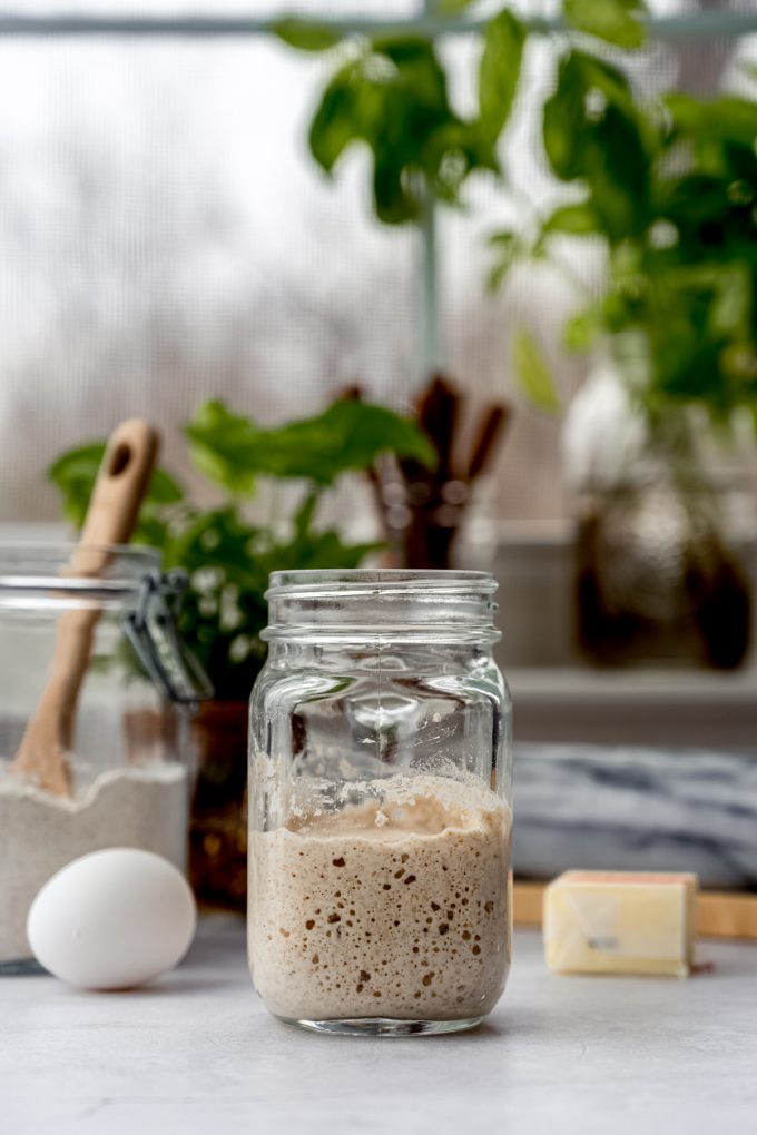 A jar of bubbly sourdough starter on a table with eggs, butter, and other ingredients around it.