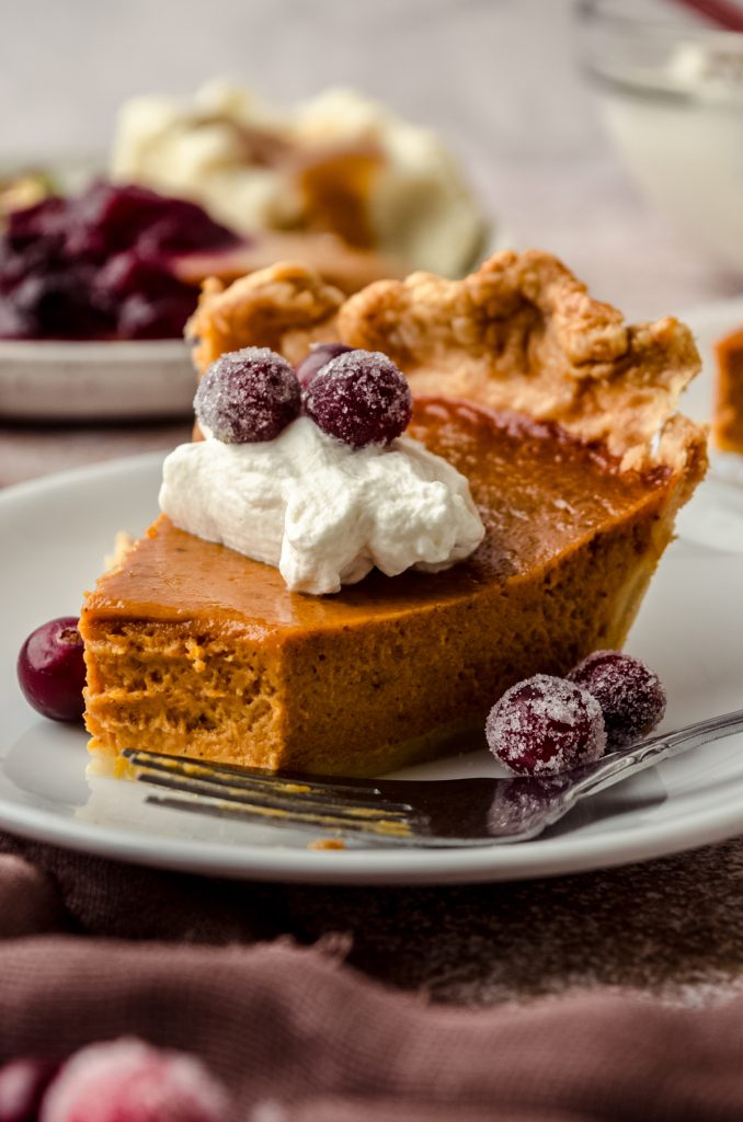 A slice of pumpkin pie on a plate with whipped cream and sugared cranberries on it as garnish and a bite has been taken out of the pie.