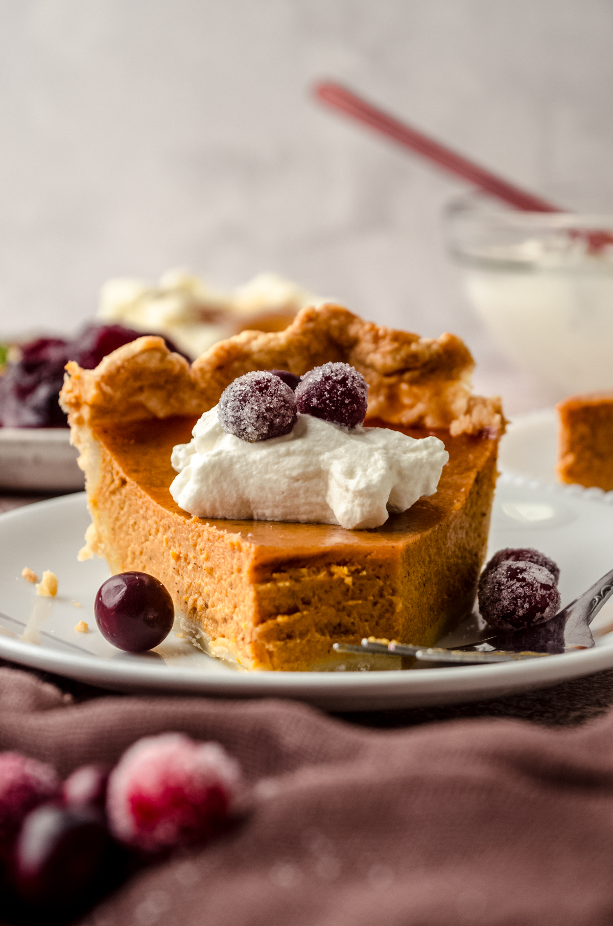 A slice of pumpkin pie on a plate with whipped cream and sugared cranberries on it as garnish and a bite has been taken out of the pie.