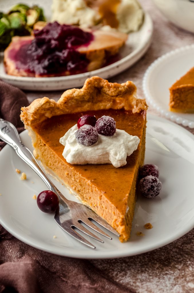 A slice of pumpkin pie on a plate with whipped cream and sugared cranberries on it as garnish.