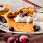 A slice of pumpkin pie on a plate with whipped cream and sugared cranberries on it as garnish.