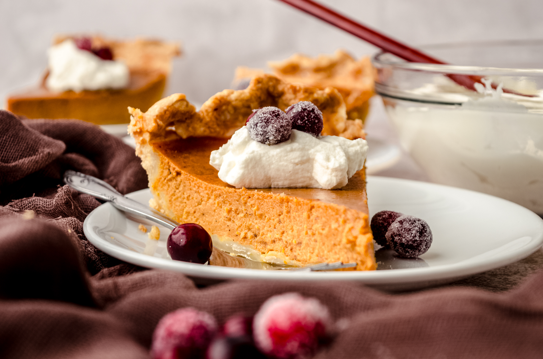 A slice of pumpkin pie on a plate with whipped cream and sugared cranberries on it as garnish.