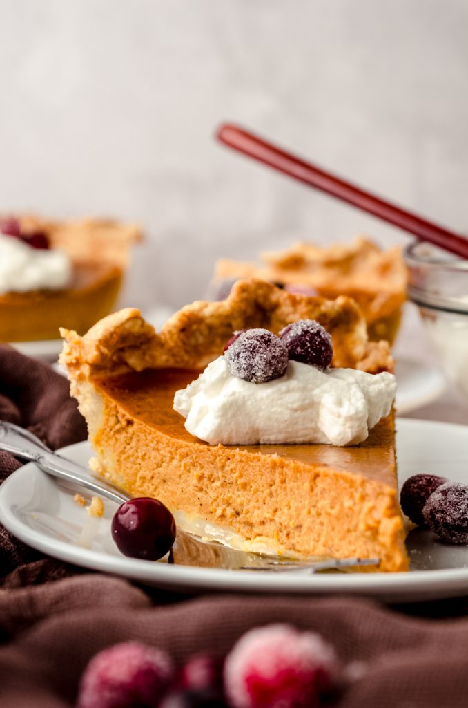 A slice of pumpkin pie on a plate with whipped cream and sugared cranberries on it as garnish.