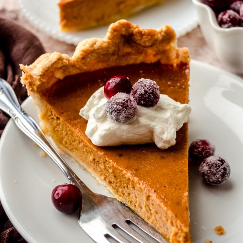 A slice of pumpkin pie on a plate with whipped cream and sugared cranberries on it as garnish.