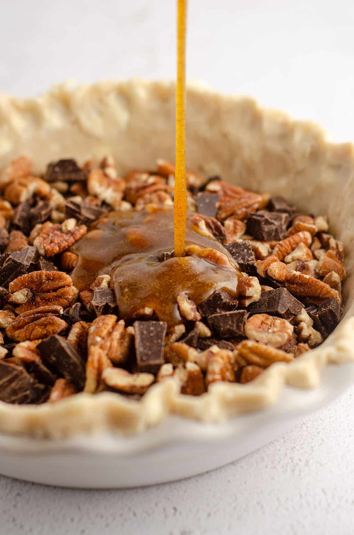 filling being poured into pie plate with a pie crust fitted into it, pie plate is filled with pecans and chocolate chunks