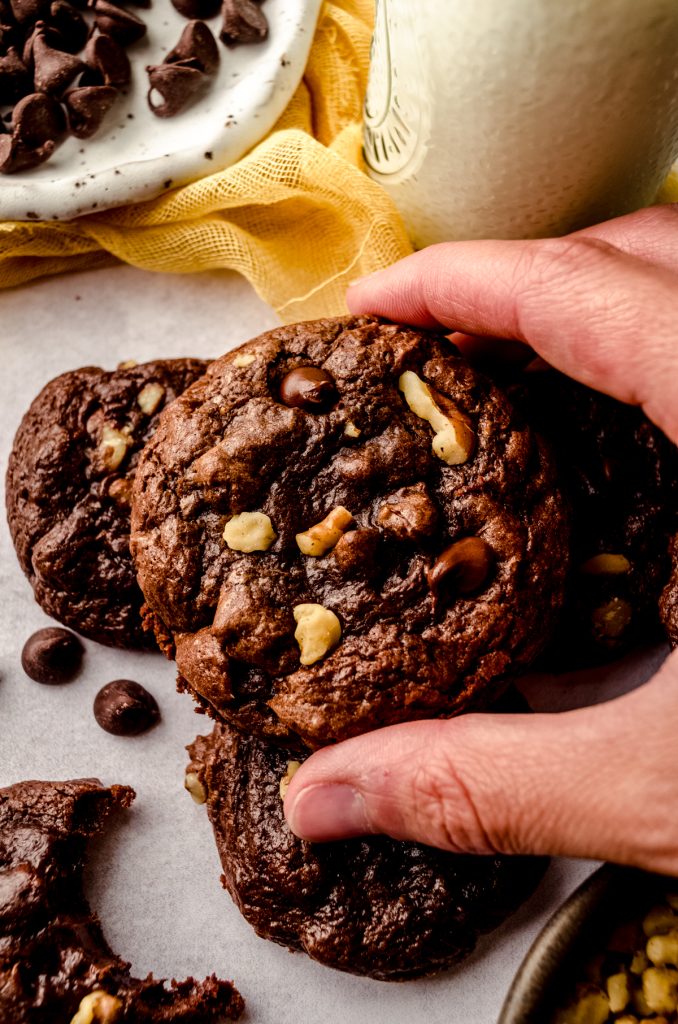 Someone is holding a brownie cookie near a yellow kitchen textile and a jug of milk.