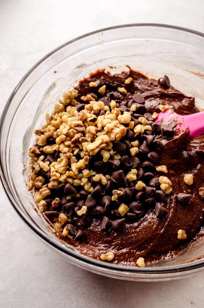 A bowl of the chocolate mixture, nuts, and chocolate chips to make brownie cookies with a pink spatula in it.