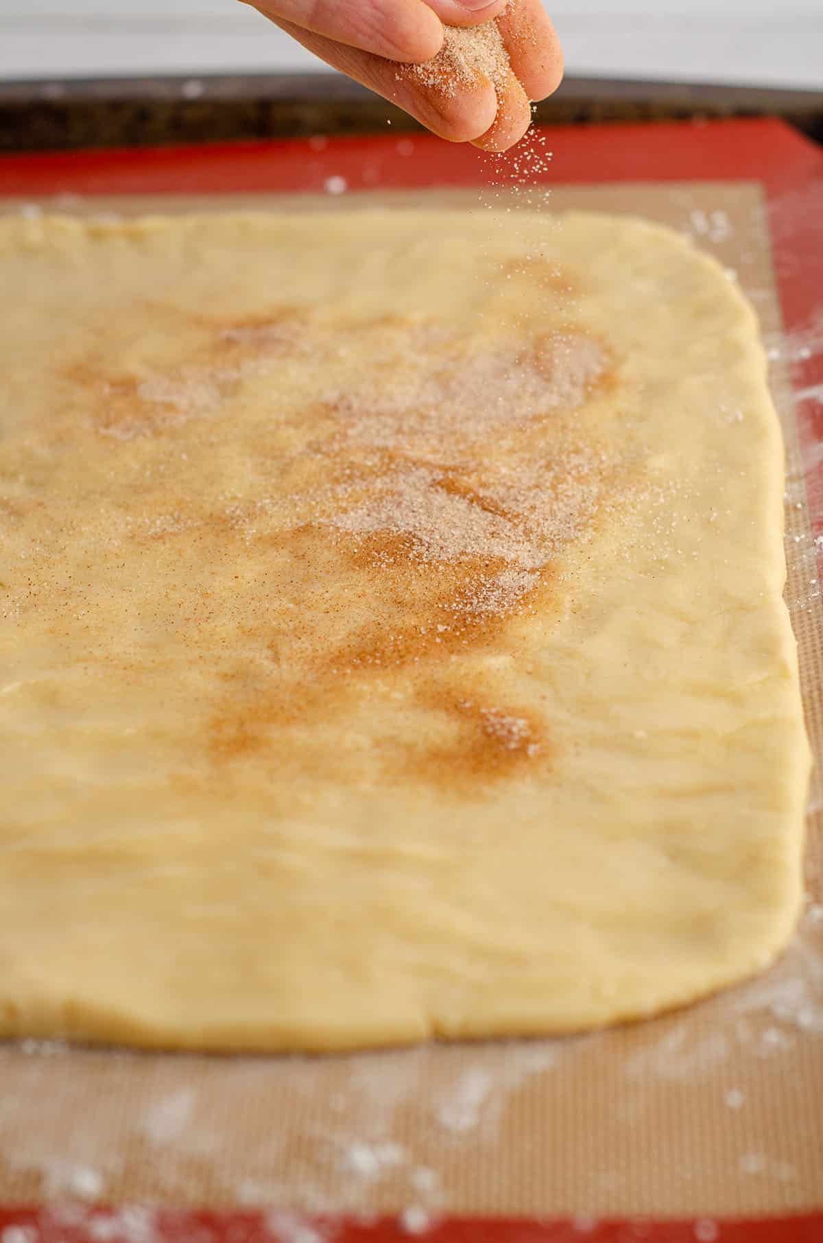 hand sprinkling cinnamon sugar on cinnamon pinwheel cookie dough rectangle