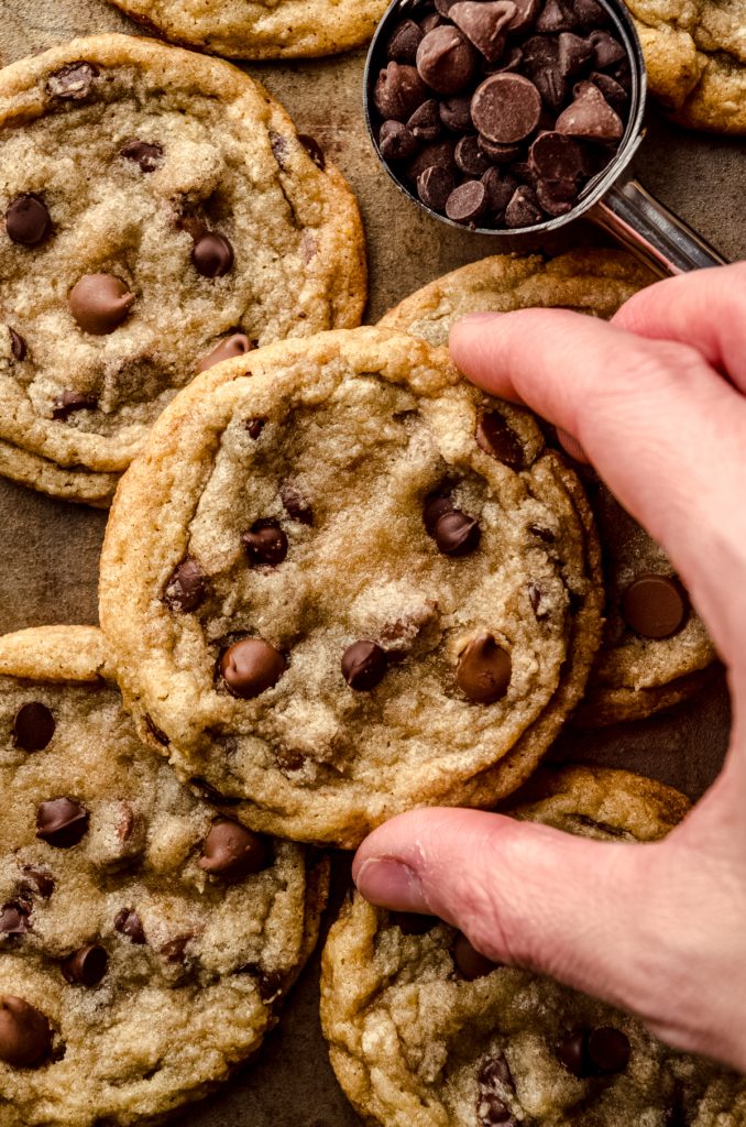 Chocolate chip cookies on a surface and someone is holding one between their thumb and pointer finger.