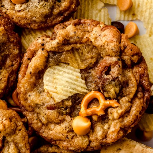 Compost cookies on a surface surrounded by pretzels, chips, and graham cracker pieces.