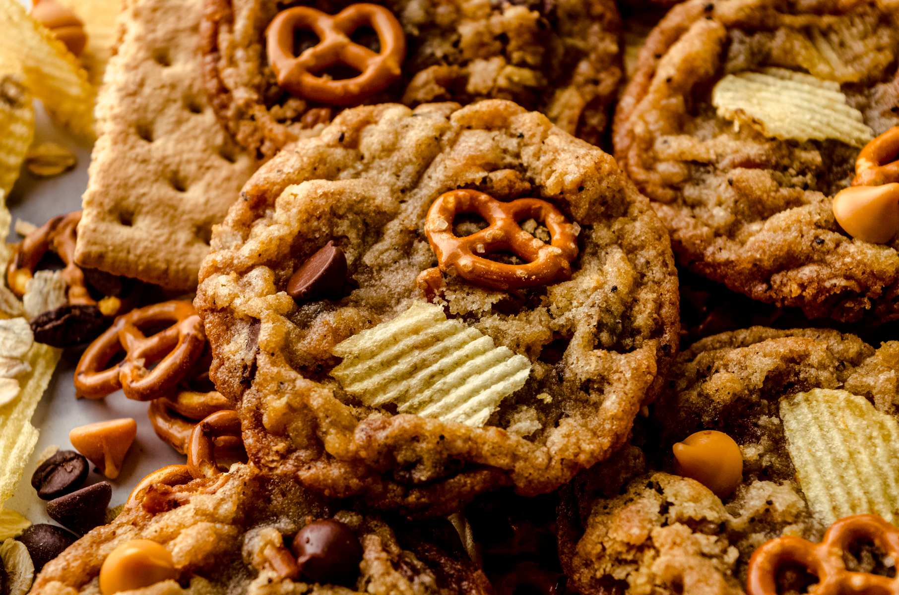 Compost cookies on a surface surrounded by pretzels, chips, and graham cracker pieces.