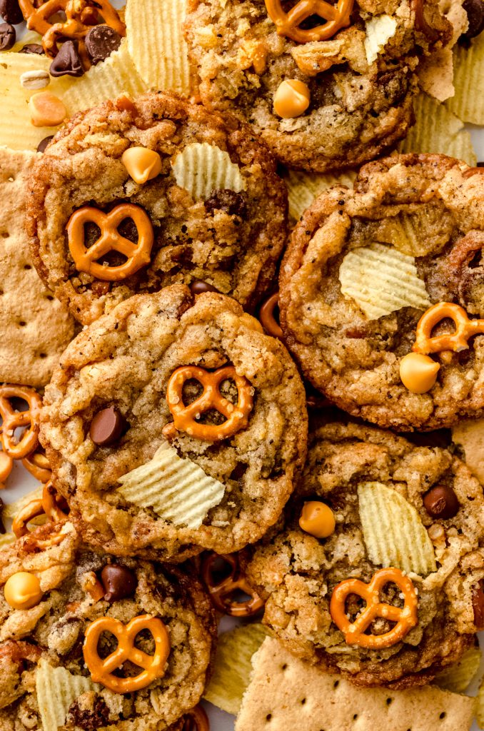 Compost cookies on a surface surrounded by pretzels, chips, and graham cracker pieces.