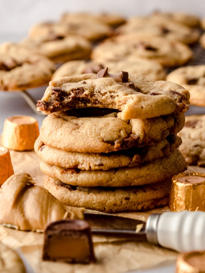 A stack of peanut butter Rolo cookies on a surface with wrapped and unwrapped Rolos around them and a bite has been taken out of the cookie on the top.