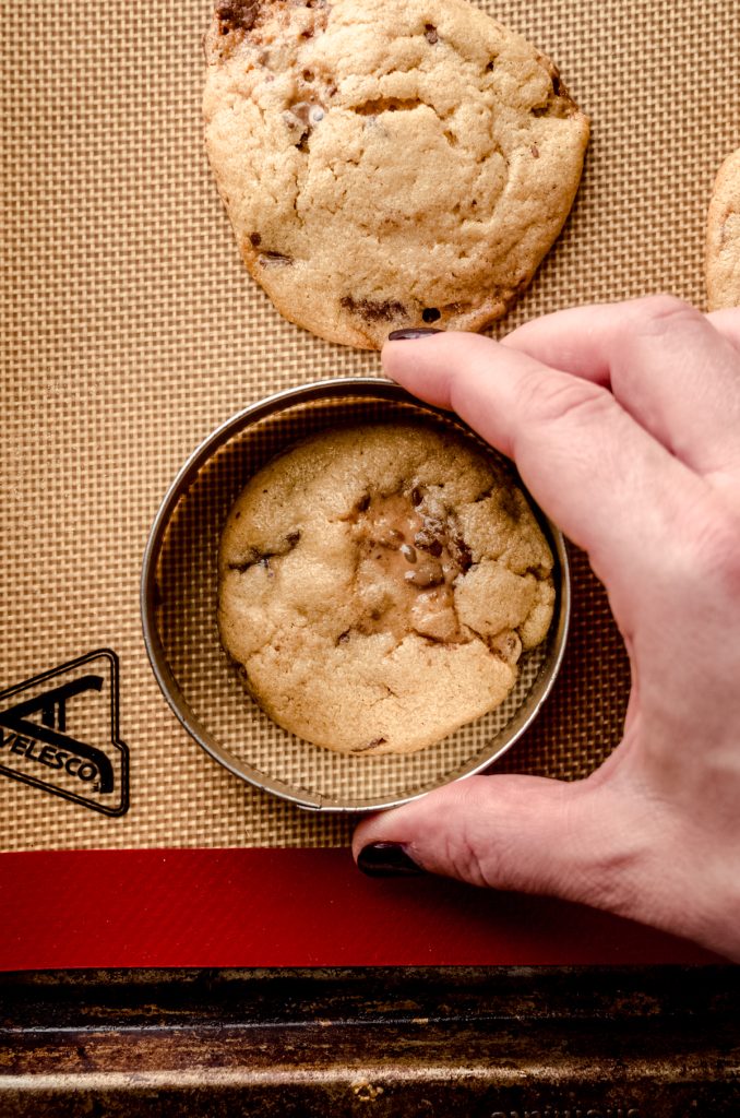 Someone is using a round cookie cutter to reshape peanut butter Rolo cookies that have just come out of the oven.