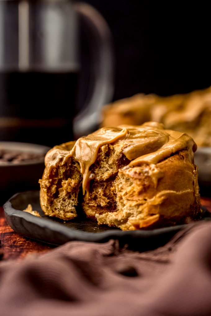 A coffee cinnamon roll on a plate with a French press in the background.