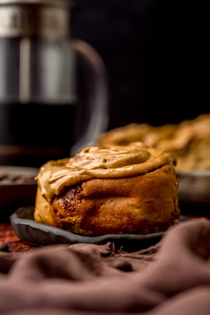 A coffee cinnamon roll on a plate with a French press in the background.