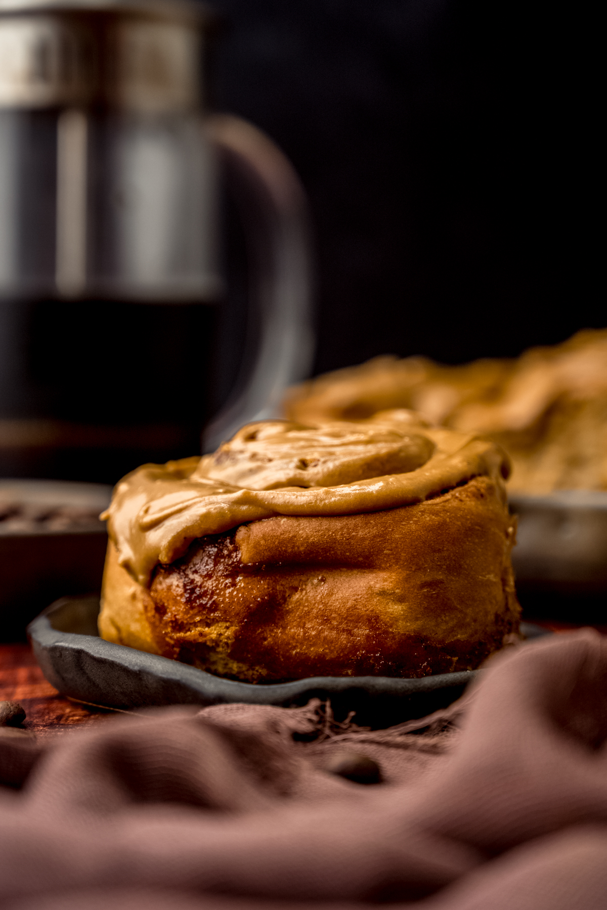 A coffee cinnamon roll on a plate with a French press in the background.
