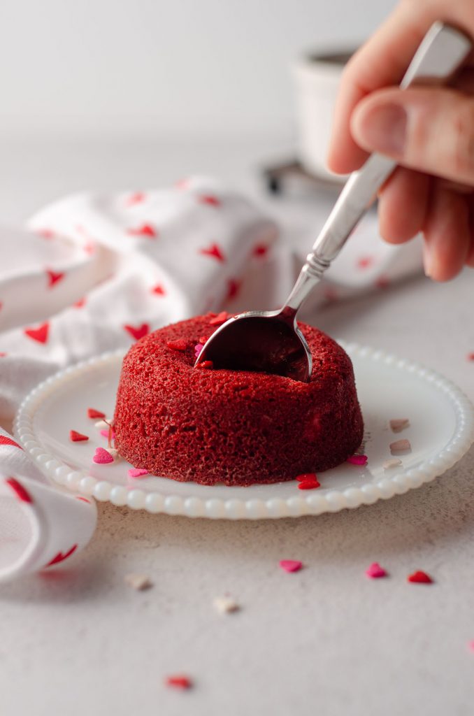 a hand with a spoon digging into a red velvet lava cake sitting on a white plate
