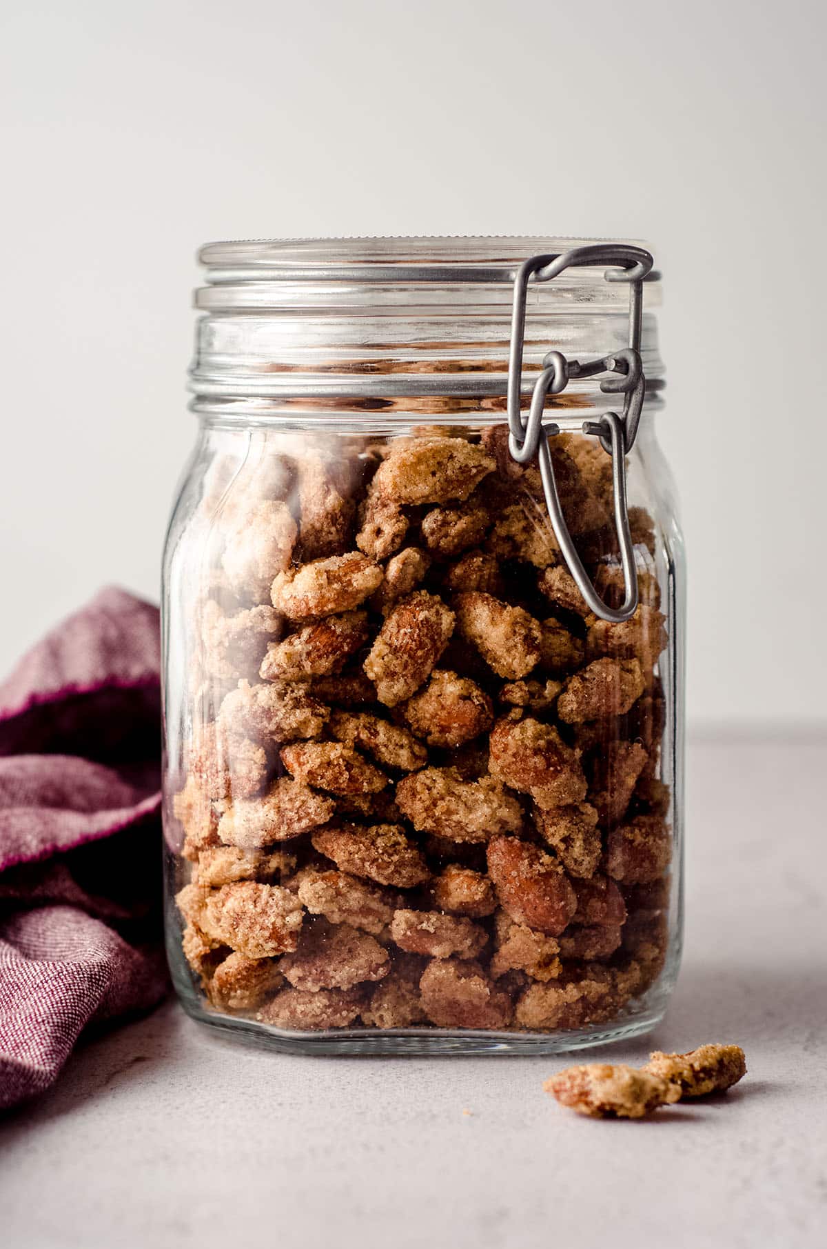 candied almonds in a jar with a plum colored kitchen towel next to it and almonds in the foreground