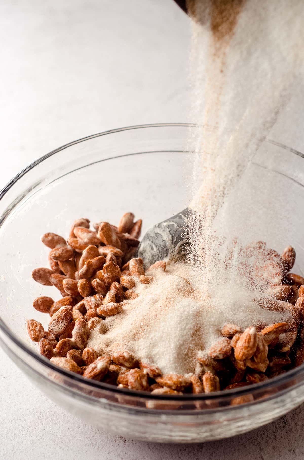 a bowl of almonds coated in vanilla bean paste and frothed egg whites with a bowl of sugar and cinnamon pouring on top to make candied almonds