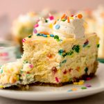 A slice of funfetti cheesecake on a plate with a fork and a bite taken out of it.