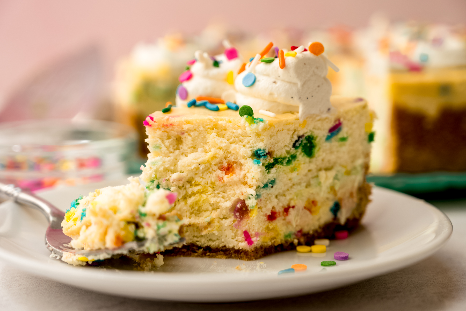 A slice of funfetti cheesecake on a plate with a fork and a bite taken out of it.