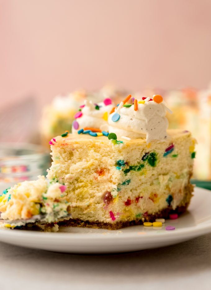 A slice of funfetti cheesecake on a plate with a fork and a bite taken out of it.