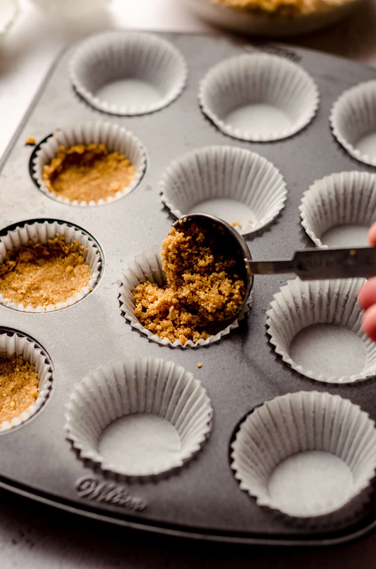 measuring spoon pouring graham cracker crust into the wells of a mini cupcake pan for no bake cheesecakes