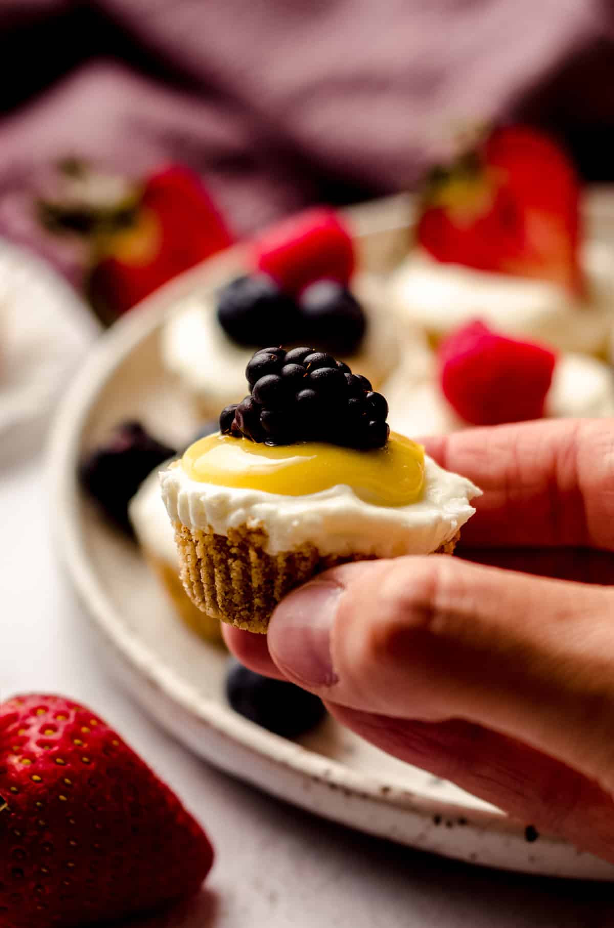 fingers holding a no bake cheesecake bite with lemon curd and a blackberry on top