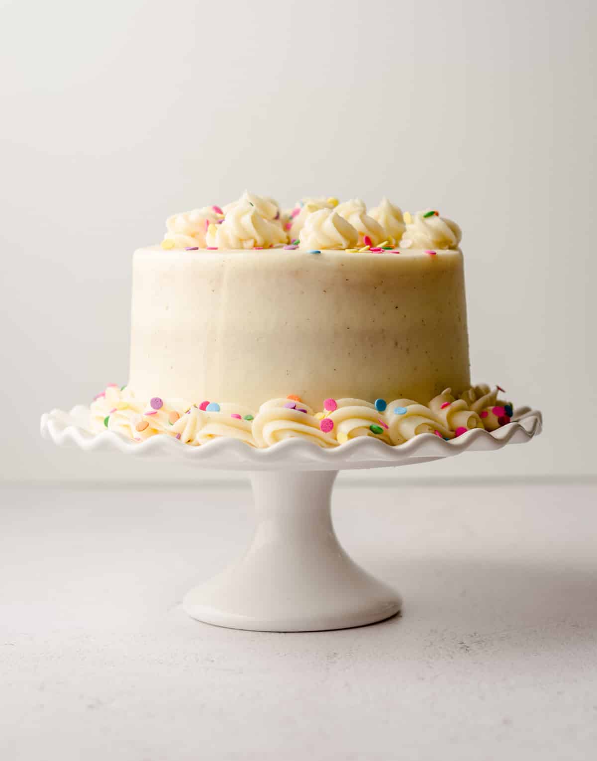 a small vanilla cake with rainbow sprinkles on a white cake stand