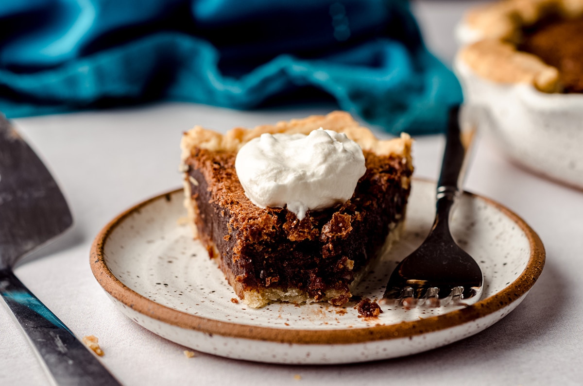 slice of chocolate chess pie on a plate with whipped cream and a fork with a bite taken out of it