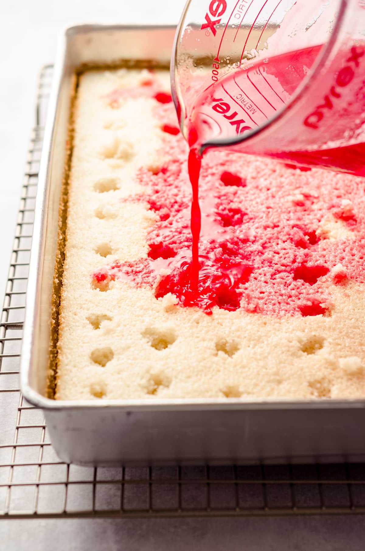 pouring strawberry jello over sheet cake to make strawberry poke cake