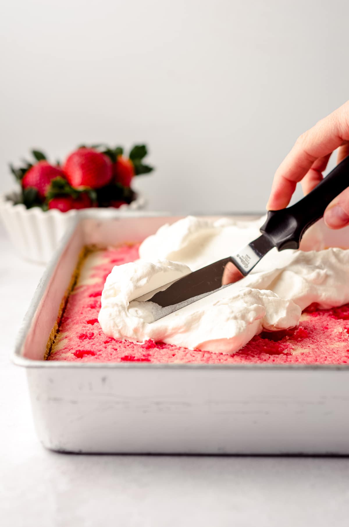 spreading whipped cream on top of strawberry poke cake in a sheet pan