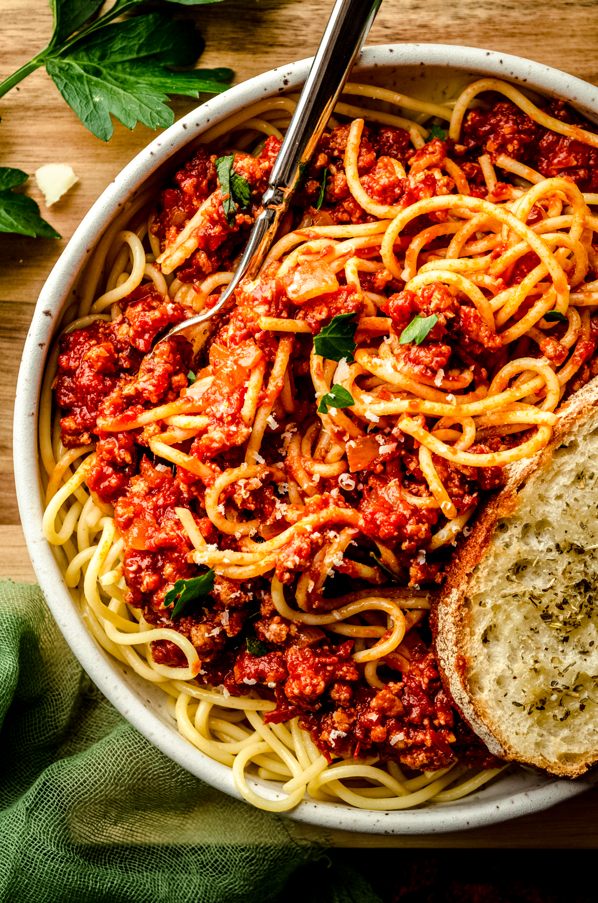 Aerial photo of a bowl of pasta with homemade meat sauce on it. There is a fork in the bowl as well as a slice of garlic bread.