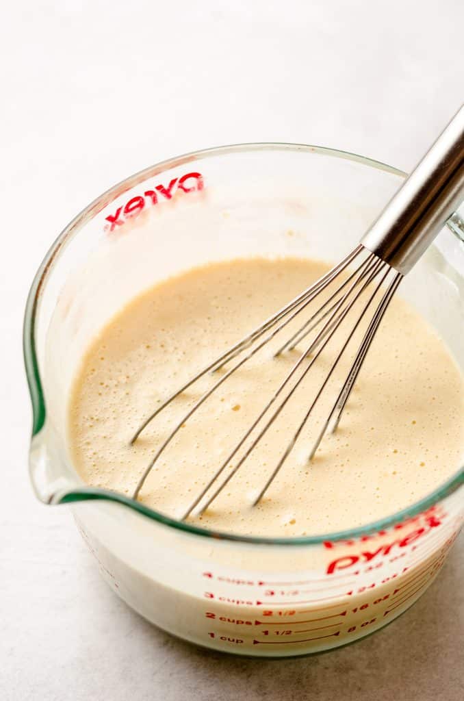 A large glass measuring cup with a crepe batter inside, being whisked.