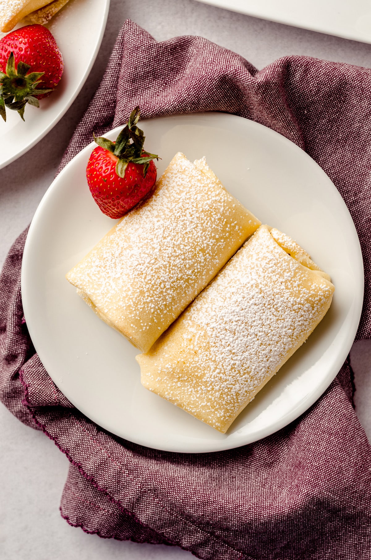 A overhead shot of a two rolled blintzes on a white plate, garnished with a single strawberry.