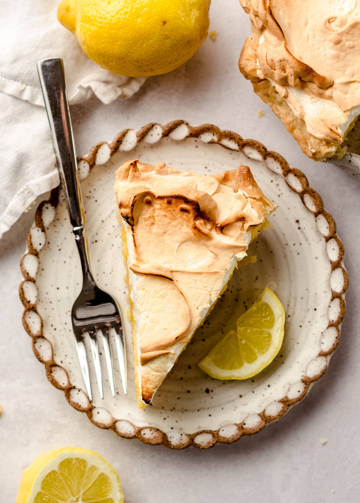 An overhead shot of a slice of lemon meringue pie with lemons on the side as garnish.