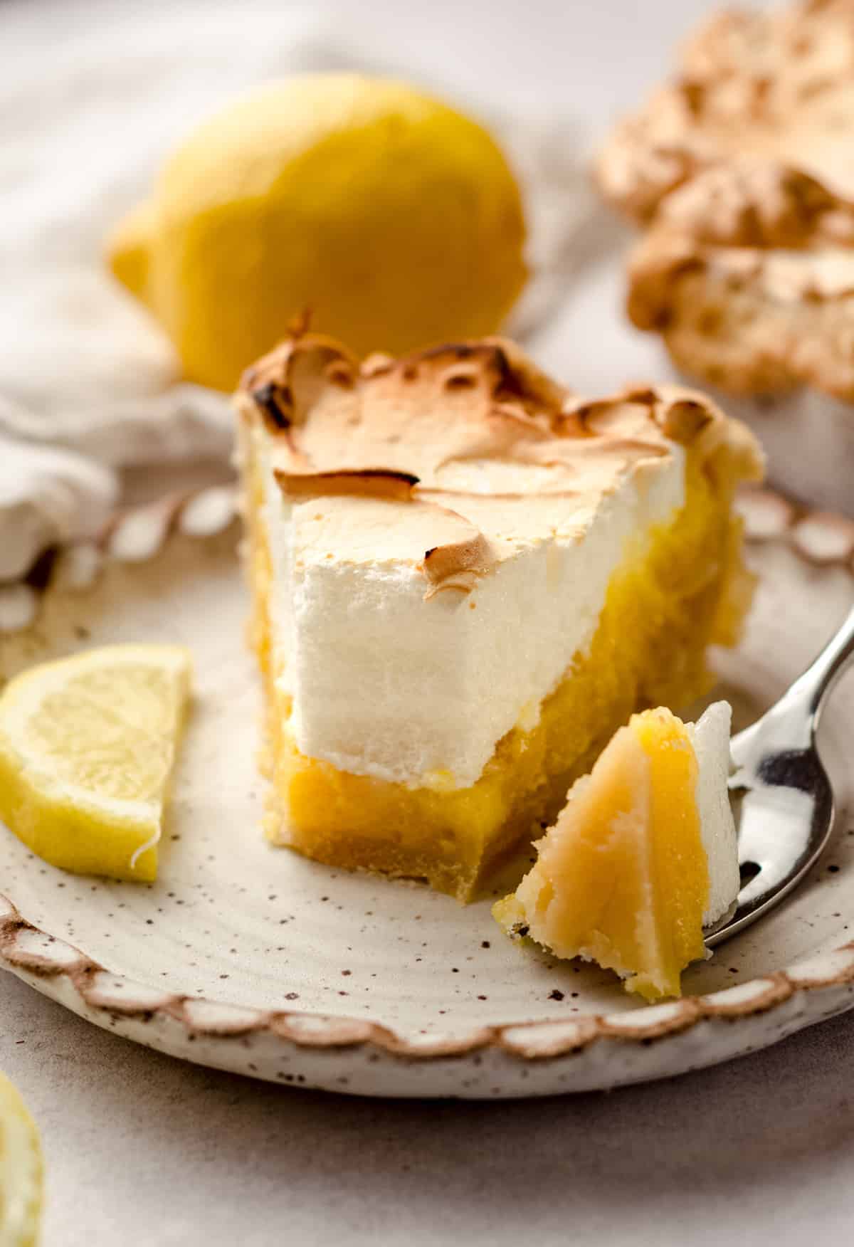 A slice of lemon meringue pie on a plate, with lemons in the background.