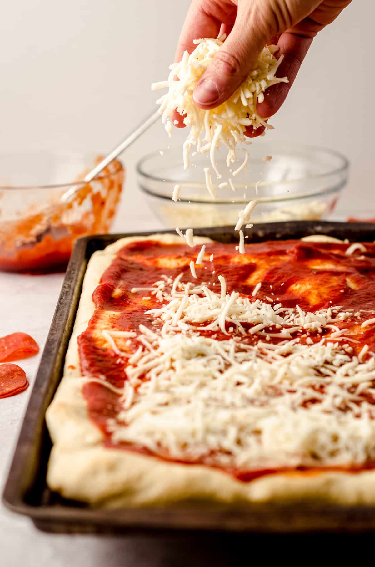 Sprinkling the cheese onto the surface of a sheet pan pizza topped with sauce.