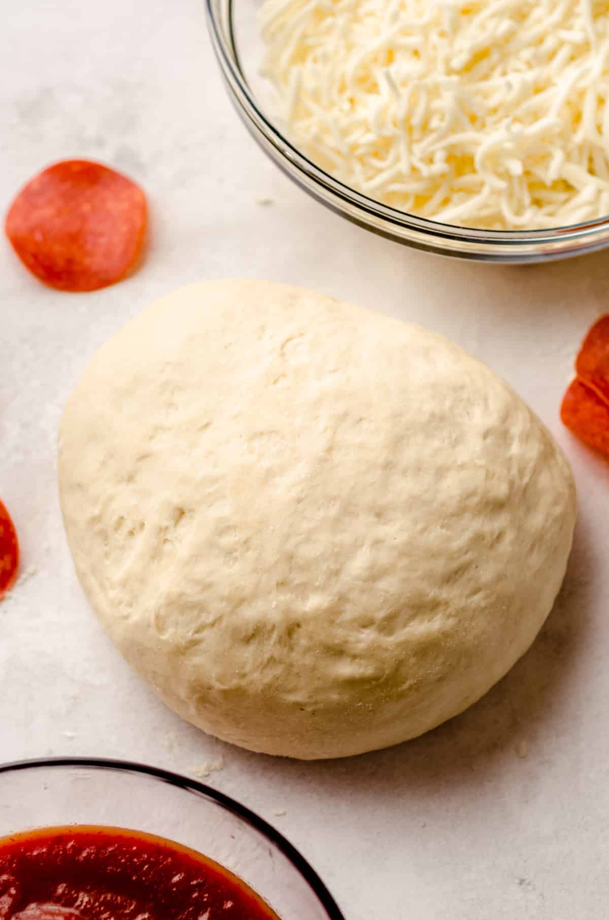 A large circle of pizza dough, with shredded mozzarella and pizza sauce in the background.
