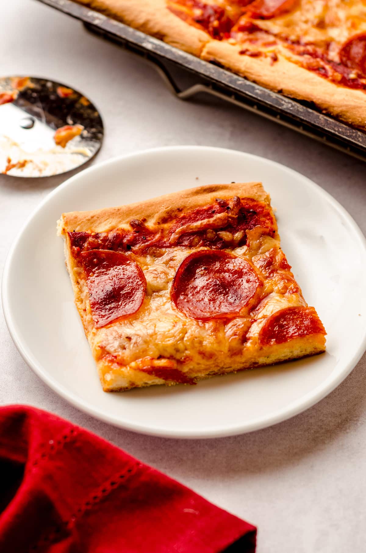 A square slice of pizza on a white plate, with a full size pizza in the background.