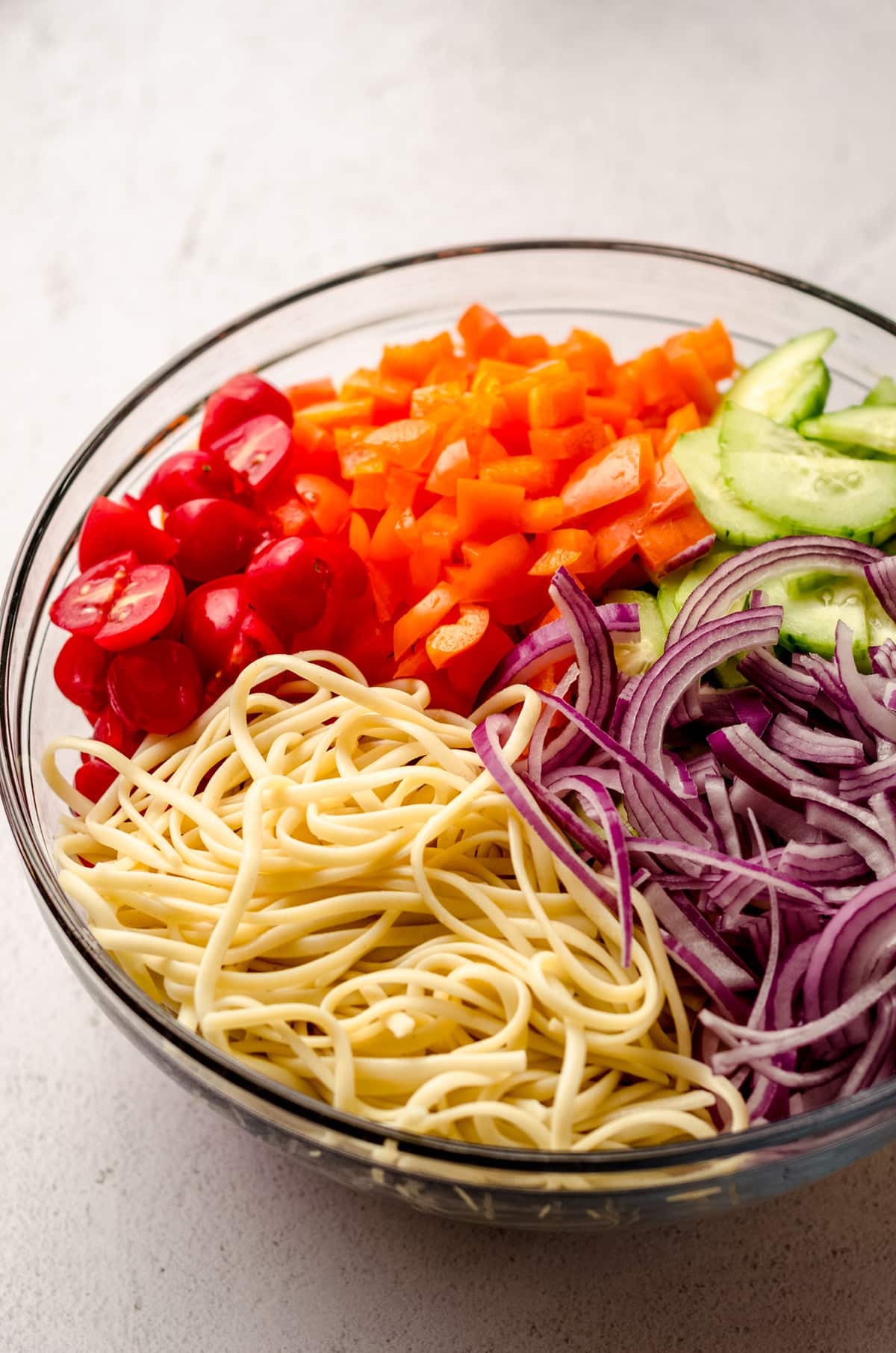 The ingredients needed to make spaghetti salad, all in a large bowl: spaghetti noodles, onion, cucumber, carrot, and bell pepper.