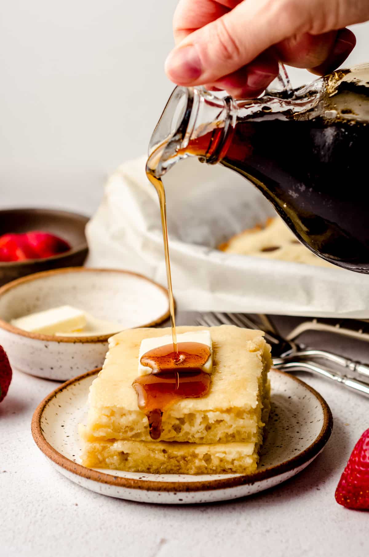 pouring syrup onto a stack of sheet pan pancakes with a pat of butter on top
