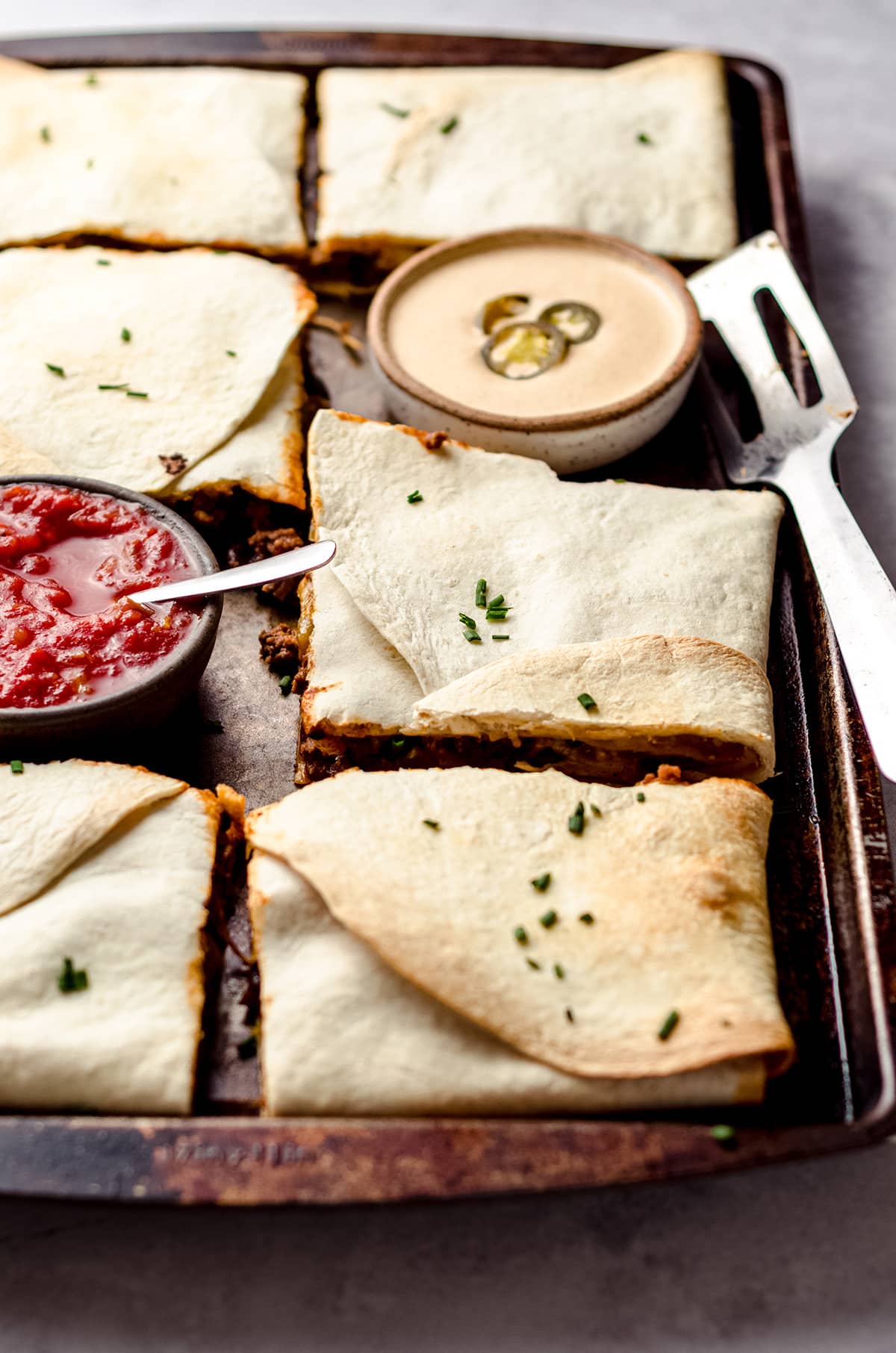A sheet pan filled with square quesadillas, and small bowls of queso and salsa.
