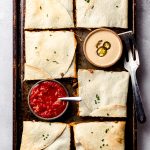 An overhead shot of a sheet pan filled with square quesadillas and served with bowls of sauce and salsa.