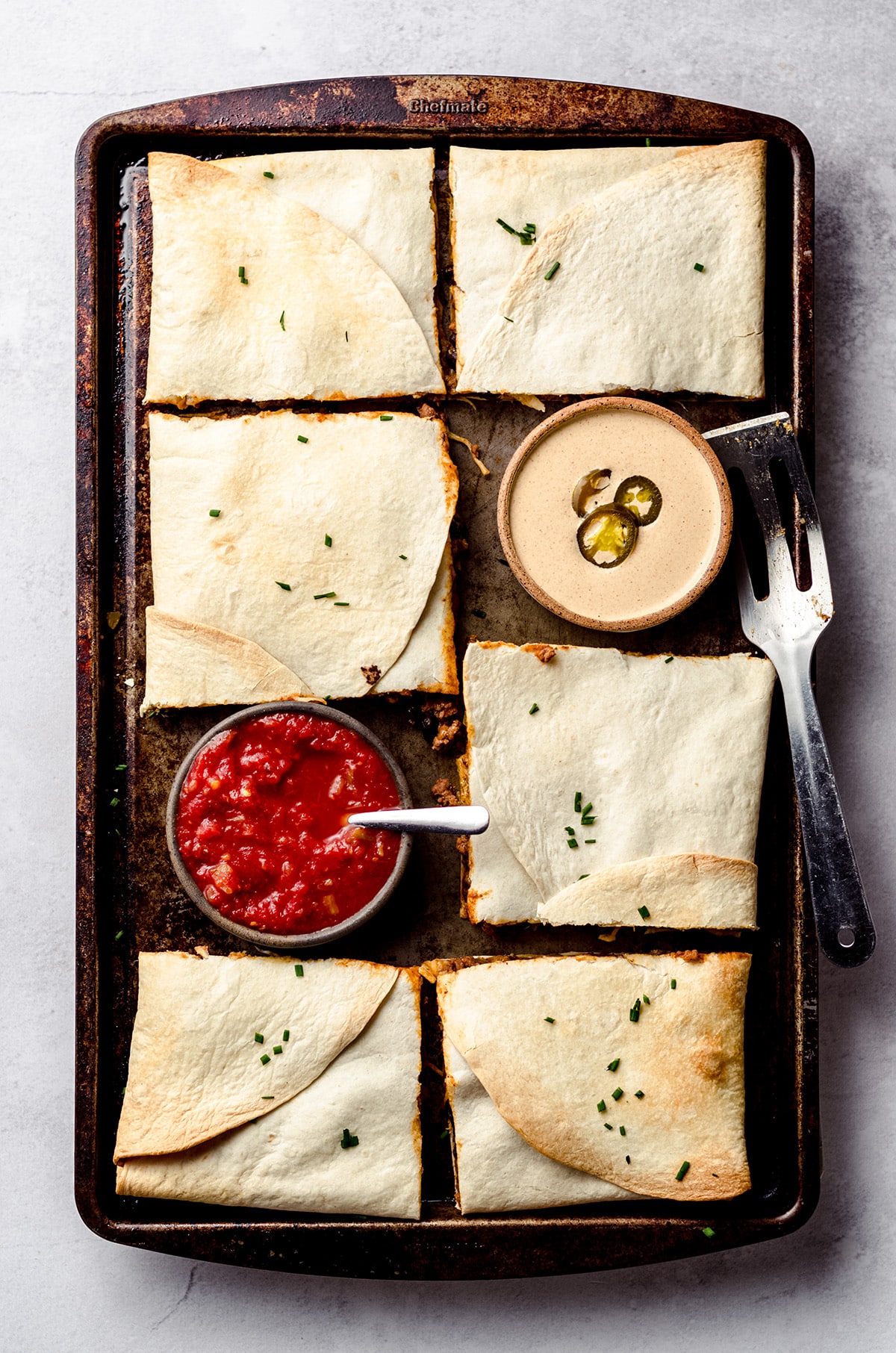 An overhead shot of a sheet pan filled with square quesadillas and served with bowls of sauce and salsa.