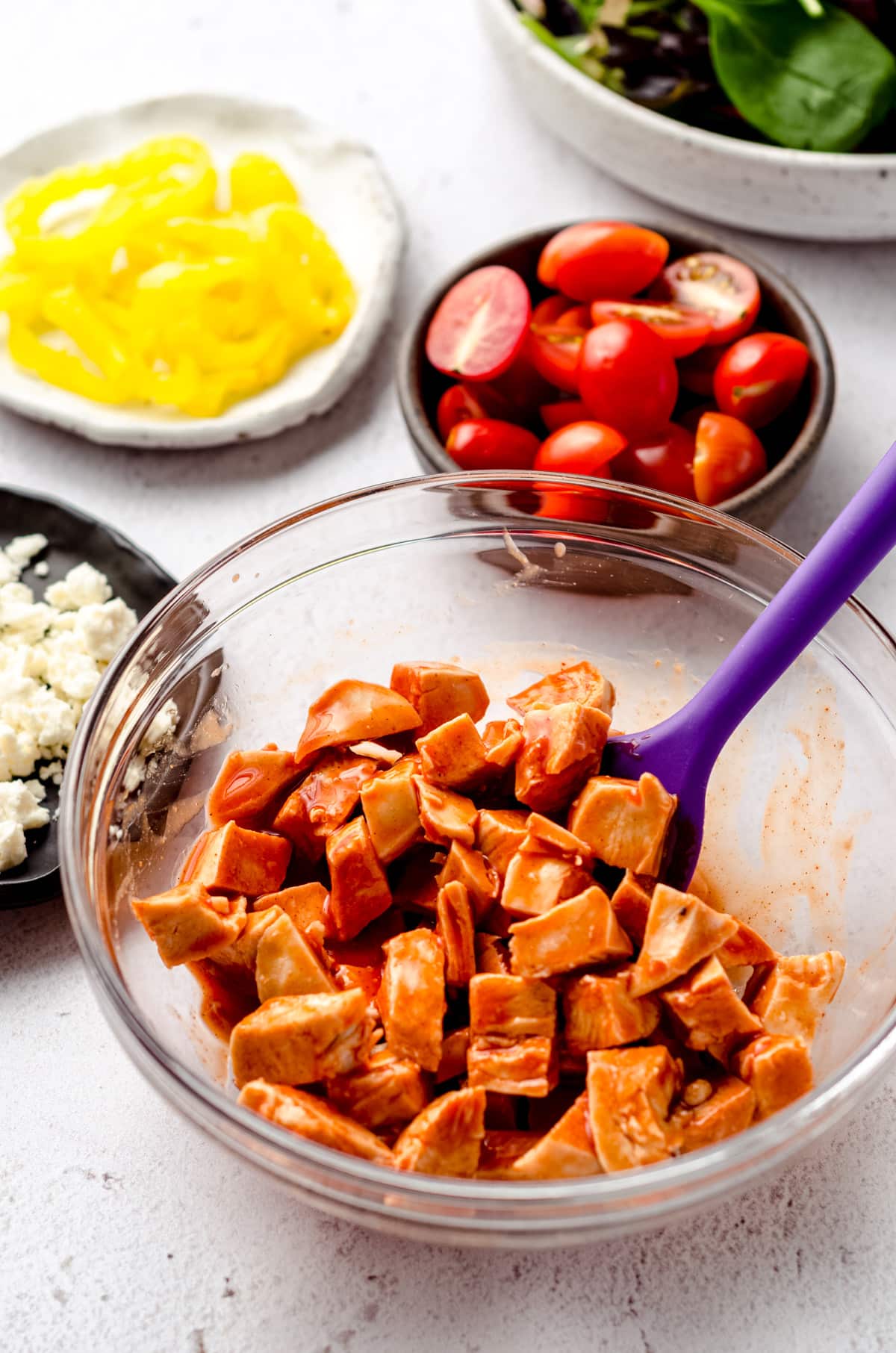 Cubed chicken in a bowl with buffalo sauce. The mixture has a purple spatula sitting in it and there are tomatoes, feta cheese, banana peppers, and mixed greens in the background.