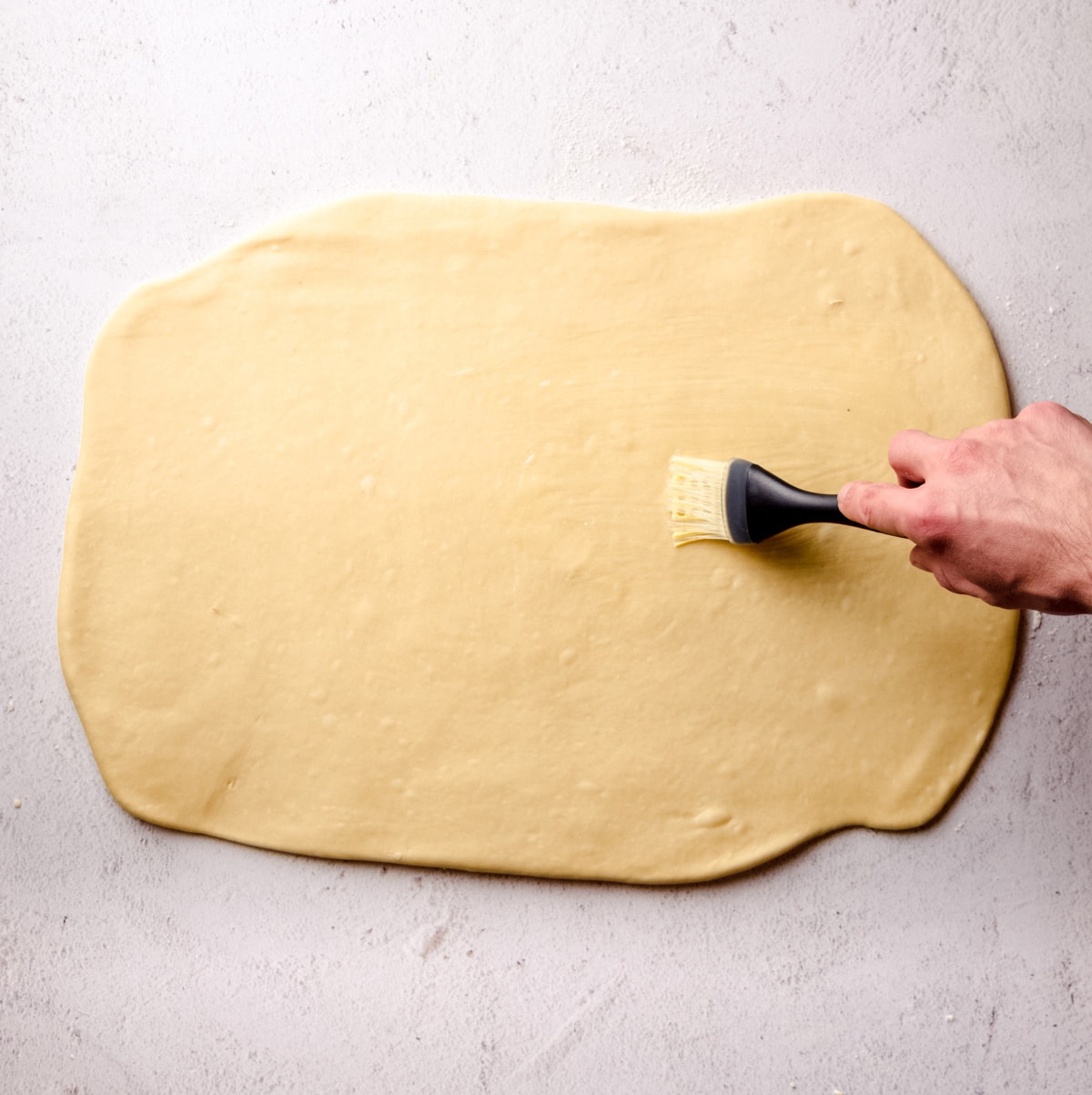 A hand brushing melted butter onto a rectangle of yeast dough for cinnamon rolls.